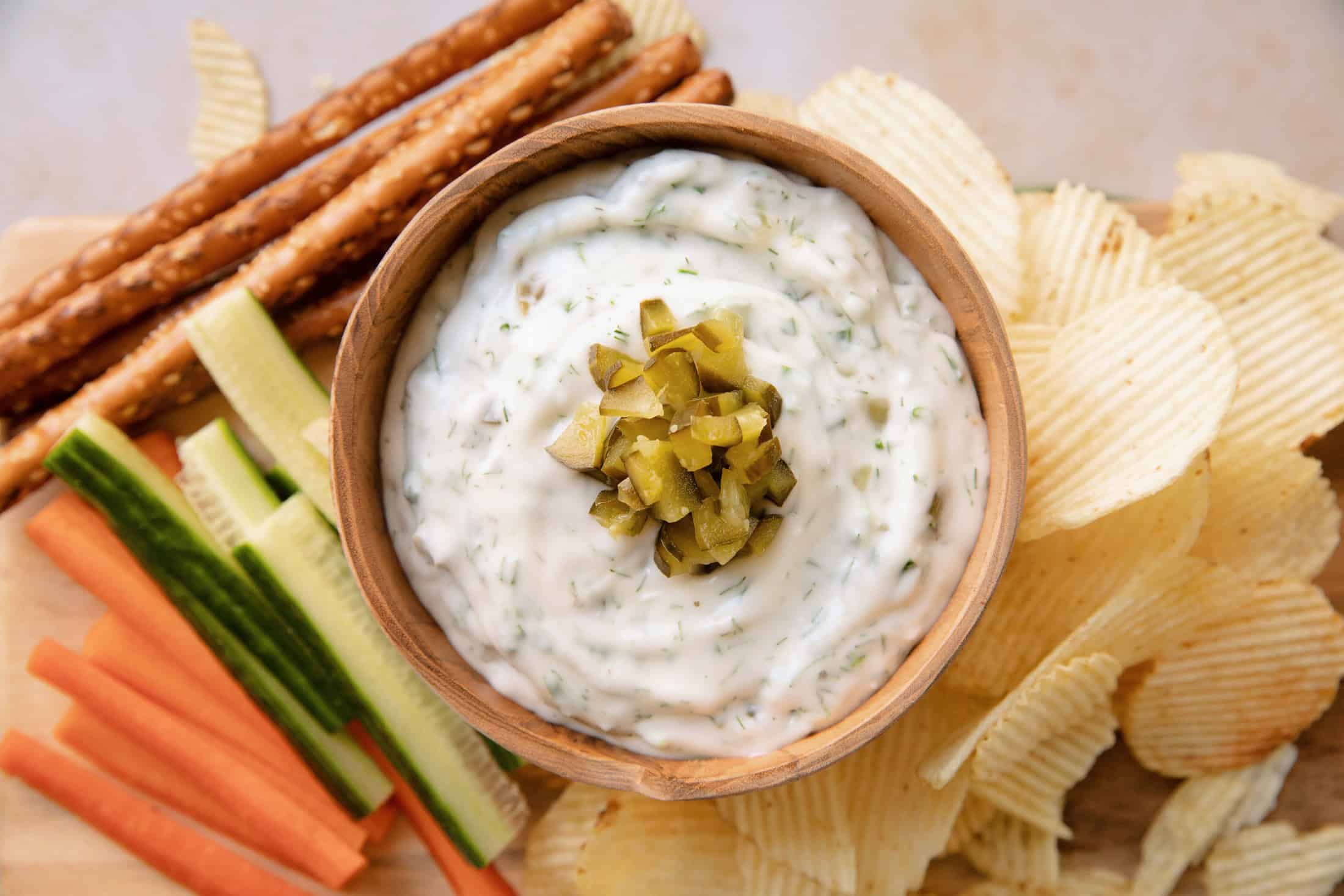 overhead of a bowl of dill pickle dip with chopped pickles on top, sitting on a tray with vegetables and chips